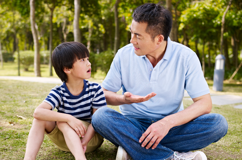 asian-father-and-elementary-age-son-sitting-on-grass-outdoors-having-a-serious-conversation